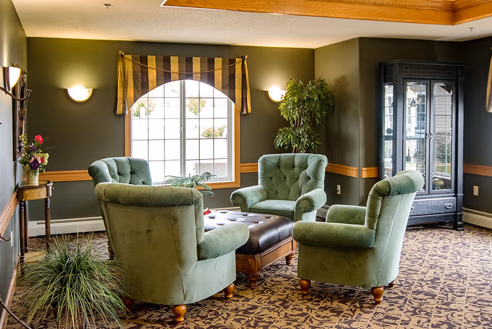 Cozy seating area with four green tufted armchairs arranged around a central leather-topped ottoman in a warmly decorated common room.