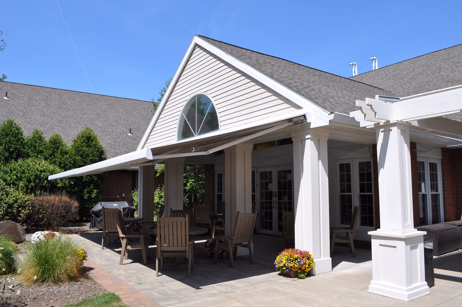 Outdoor patio area at Chapel Oaks with wooden chairs and tables under a white awning attached to a building with large windows and a triangular window above. There are plants and flowers around the patio and a clear blue sky overhead.