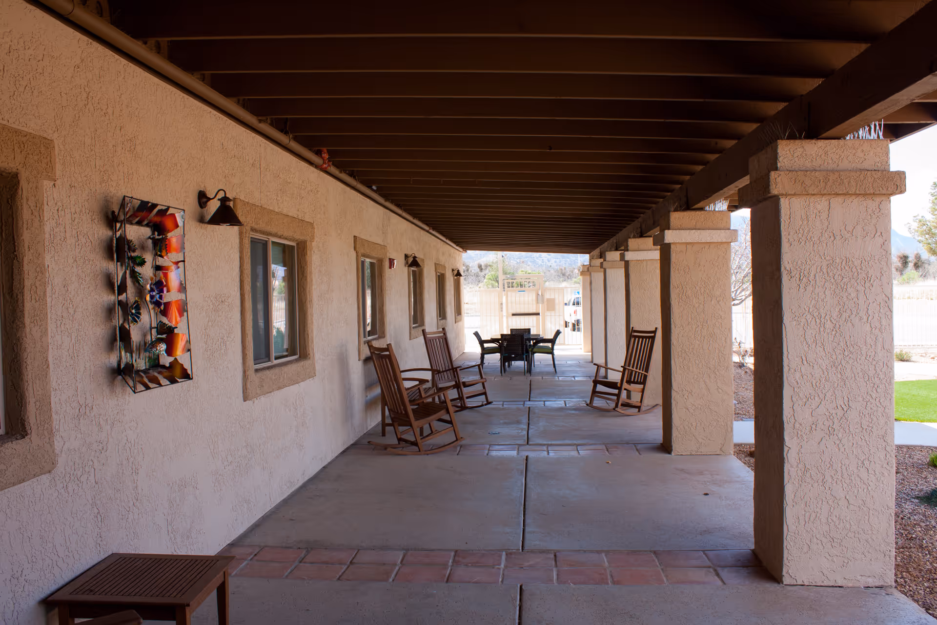 Covered outdoor patio area with several wooden rocking chairs along a textured wall with windows and decorative wall art, supported by large square columns, and a table with chairs at the far end under the roof.