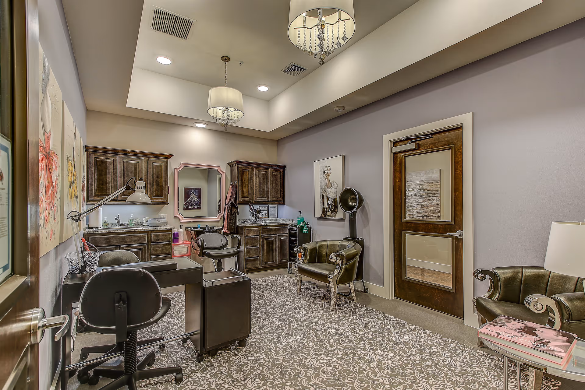 Interior view of a well-lit salon or beauty room with two salon chairs, a hair dryer, wooden cabinets, a mirror, and comfortable seating including two armchairs. The room has patterned carpet flooring, soft lighting from ceiling fixtures, and decorative artwork on the walls.