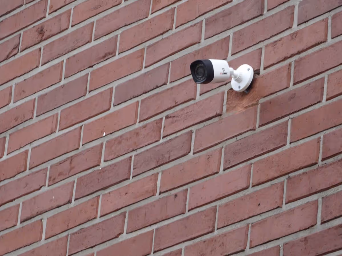 A white security camera mounted on a red brick wall, angled slightly downward.