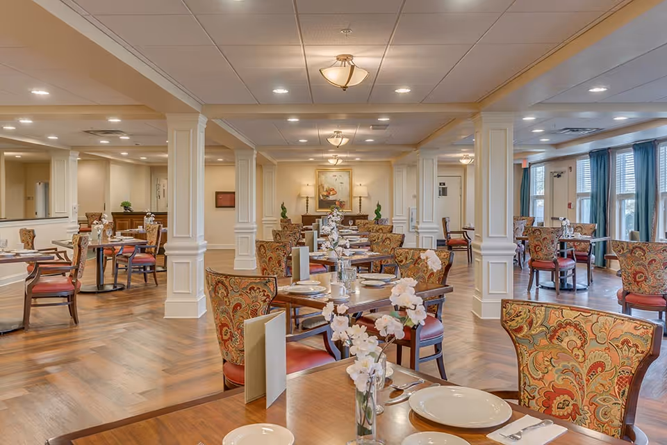 A spacious and elegant dining room with multiple tables set with plates, cups, and silverware. The chairs have ornate patterned upholstery and the room features white columns, wooden flooring, and soft lighting from ceiling fixtures. Large windows with blue curtains allow natural light to enter the room.