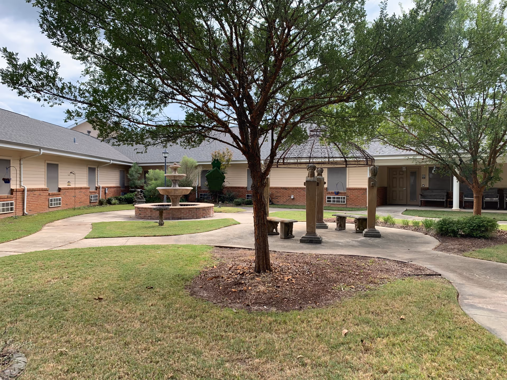 Outdoor courtyard area at Magnolia Assisted Living featuring a central fountain, paved walkways, green grass, trees, and a shaded seating area with benches and stone pillars.