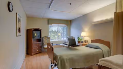 Bedroom in a senior facility with a bed, dresser, desk, chair, and window.