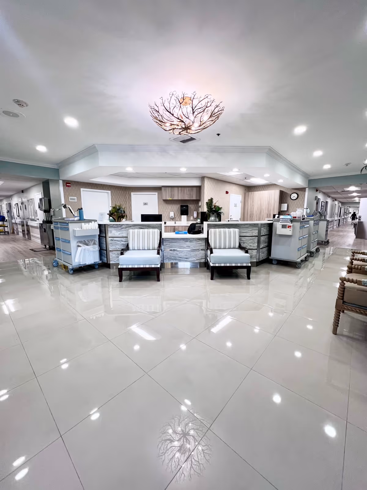 A clean and well-lit nursing station area in a senior living facility with two striped armchairs in front of the desk, medical carts on either side, and a decorative ceiling light fixture reflecting on the shiny tiled floor. Hallways extend on both sides of the station.