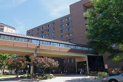 Exterior view of a multi-story brick senior living facility with a covered walkway connecting two buildings. Trees and landscaping are visible around the entrance area, with a few people walking nearby.