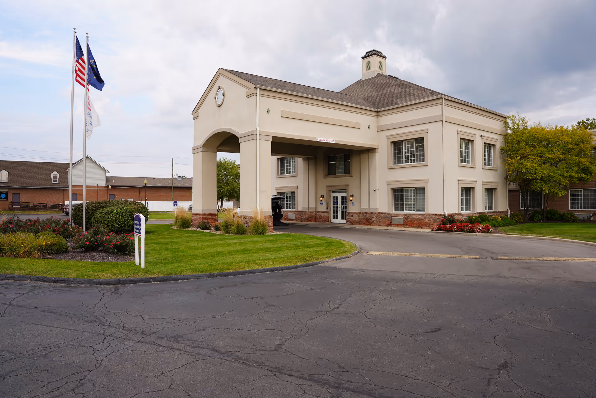 Exterior view of a senior living facility building with a covered entrance, manicured lawn, flower beds, and three flagpoles displaying the American flag, a state flag, and another flag. The sky is cloudy.
