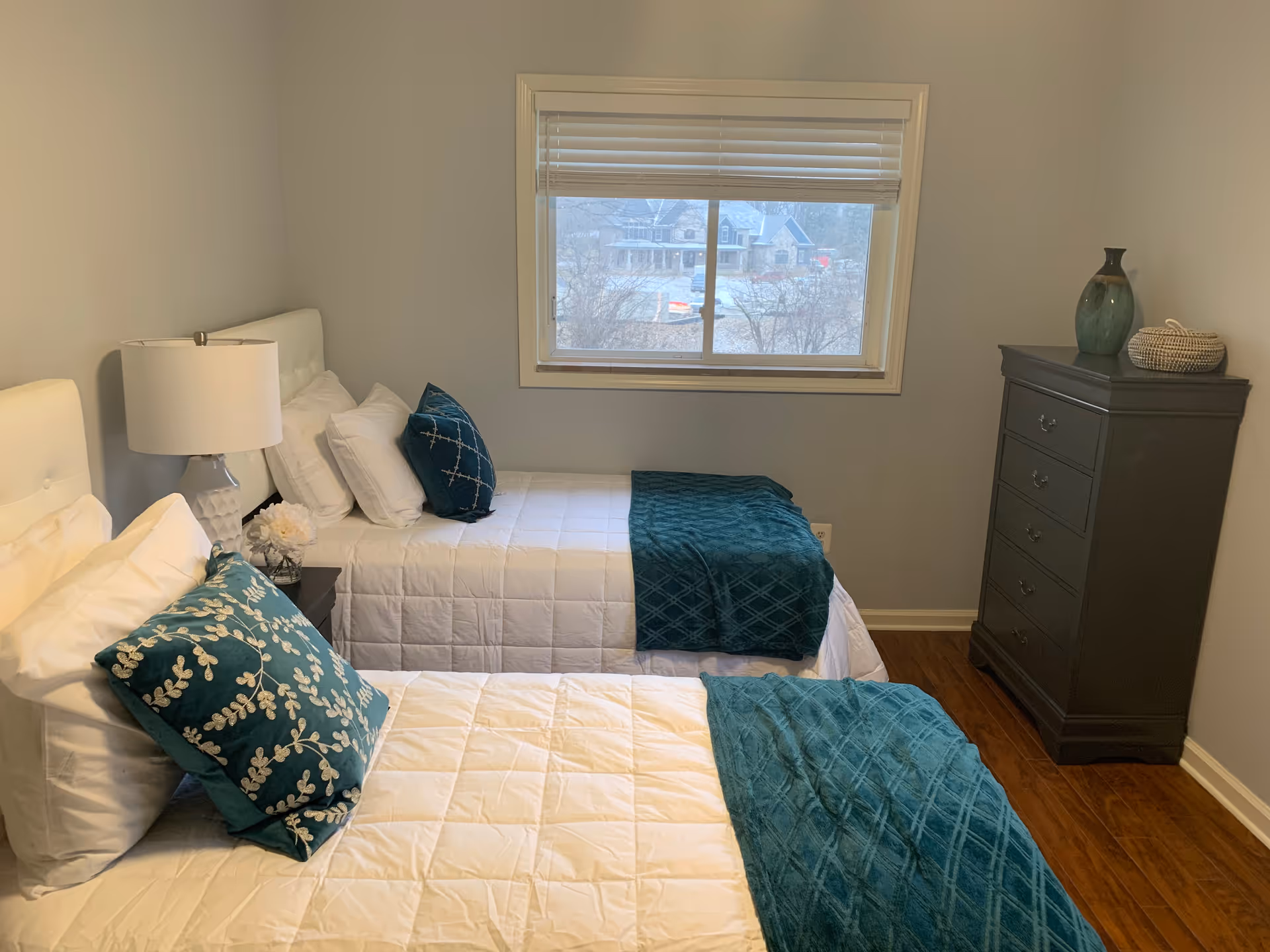 A bedroom with two twin beds, each made with white bedding and teal decorative pillows and throws. There is a nightstand with a lamp and a small flower arrangement between the beds. A window with blinds is centered on the wall behind the beds, and a dark wooden dresser with decorative items is placed against the right wall. The room has light gray walls and wooden flooring.