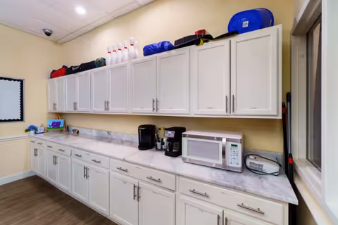 A long countertop with white cabinets above and below in a room with beige walls and wood flooring. On the countertop are a coffee maker, microwave, and a small radio. On top of the upper cabinets are various items including bowling pins and bags. There is a window on the right side and a security camera mounted on the ceiling in the corner.