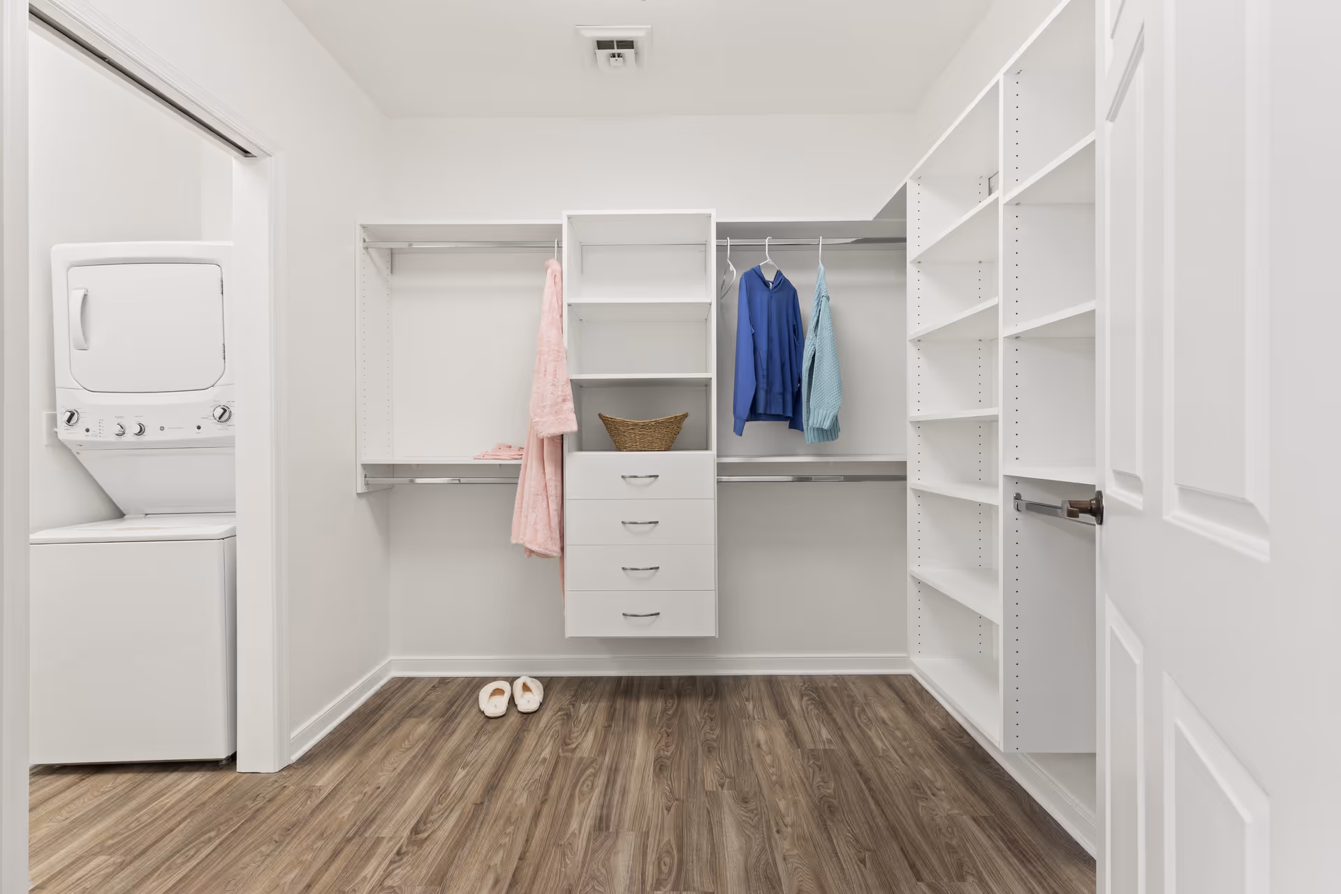 A spacious walk-in closet with white shelving units, drawers, and hanging rods. There are a few clothes hanging, including a pink robe, a blue shirt, and a light blue garment. A pair of slippers is placed on the wooden floor. To the left, there is a small laundry area with a stacked washer and dryer unit.