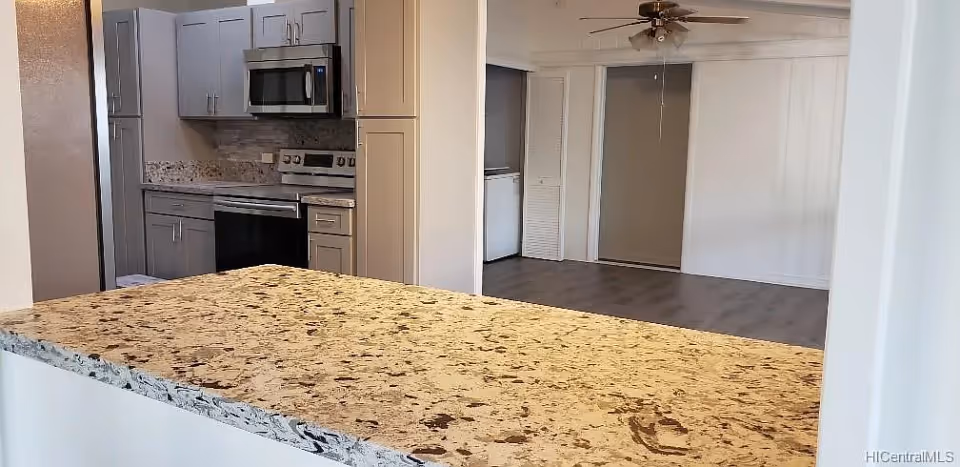 View of a modern kitchen with gray cabinets, stainless steel appliances including a microwave and stove, and a granite countertop in the foreground. The kitchen opens into an adjacent room with wood flooring, a ceiling fan, and white walls.