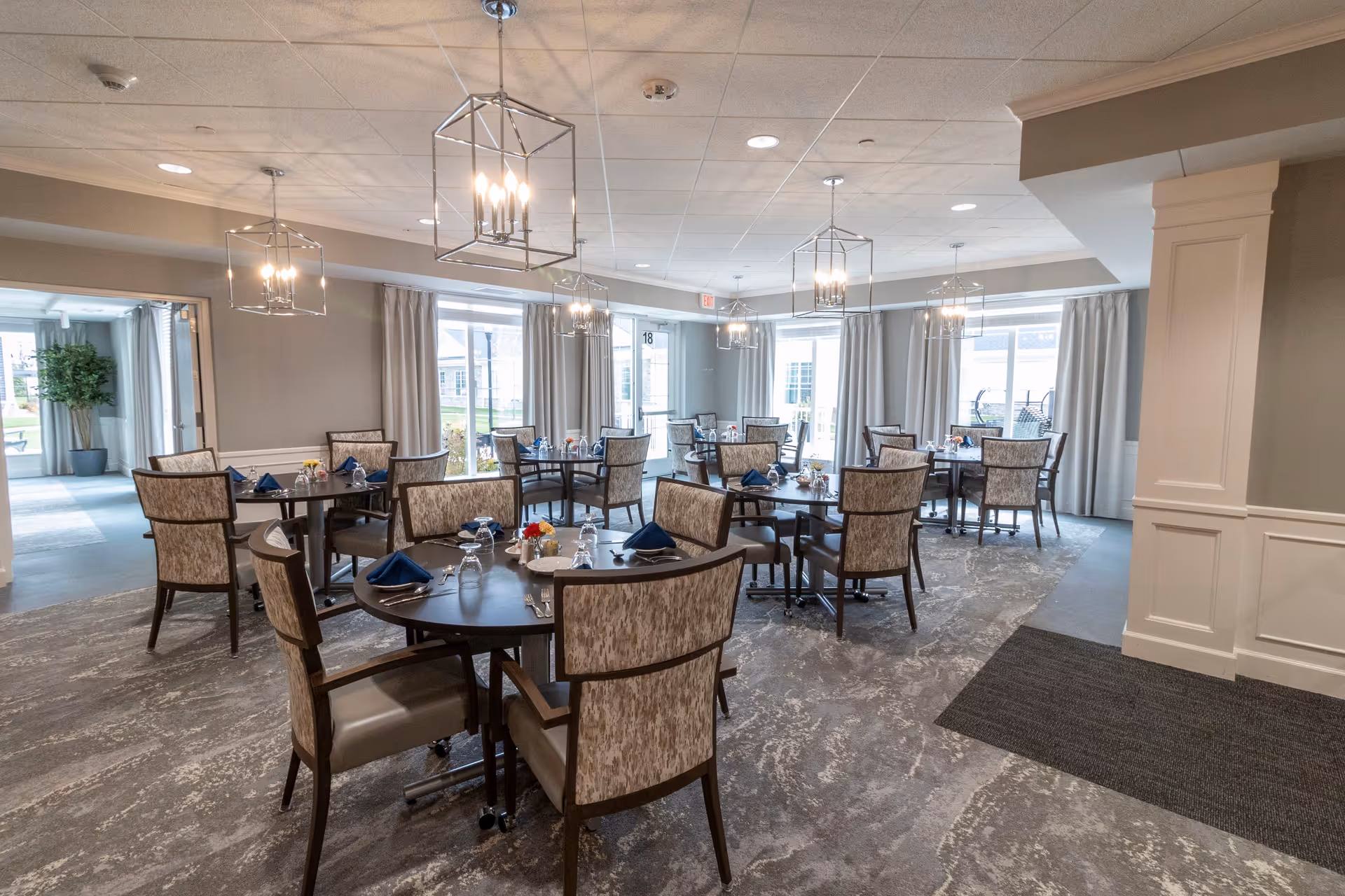 A bright and spacious dining room with multiple round tables set with blue napkins, glasses, and silverware. The room features patterned carpet, large windows with curtains allowing natural light, and modern pendant lights hanging from the ceiling. There is a doorway leading to another area with a large potted plant visible.