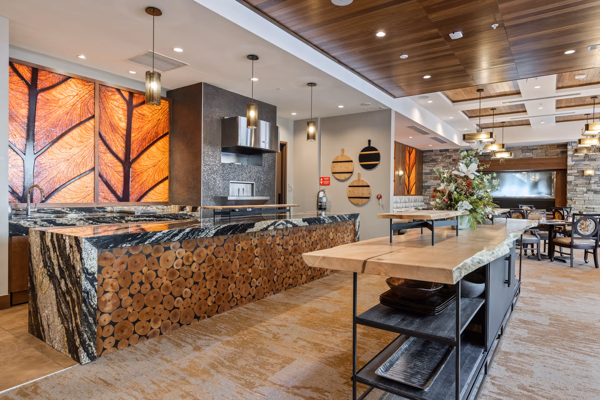 A modern and stylish dining area in Aegis Living Mercer Island featuring a large kitchen island with a unique wood log design on the side, a countertop with a natural wood finish, pendant lighting, decorative wall art with leaf patterns, and a seating area with tables and chairs in the background.