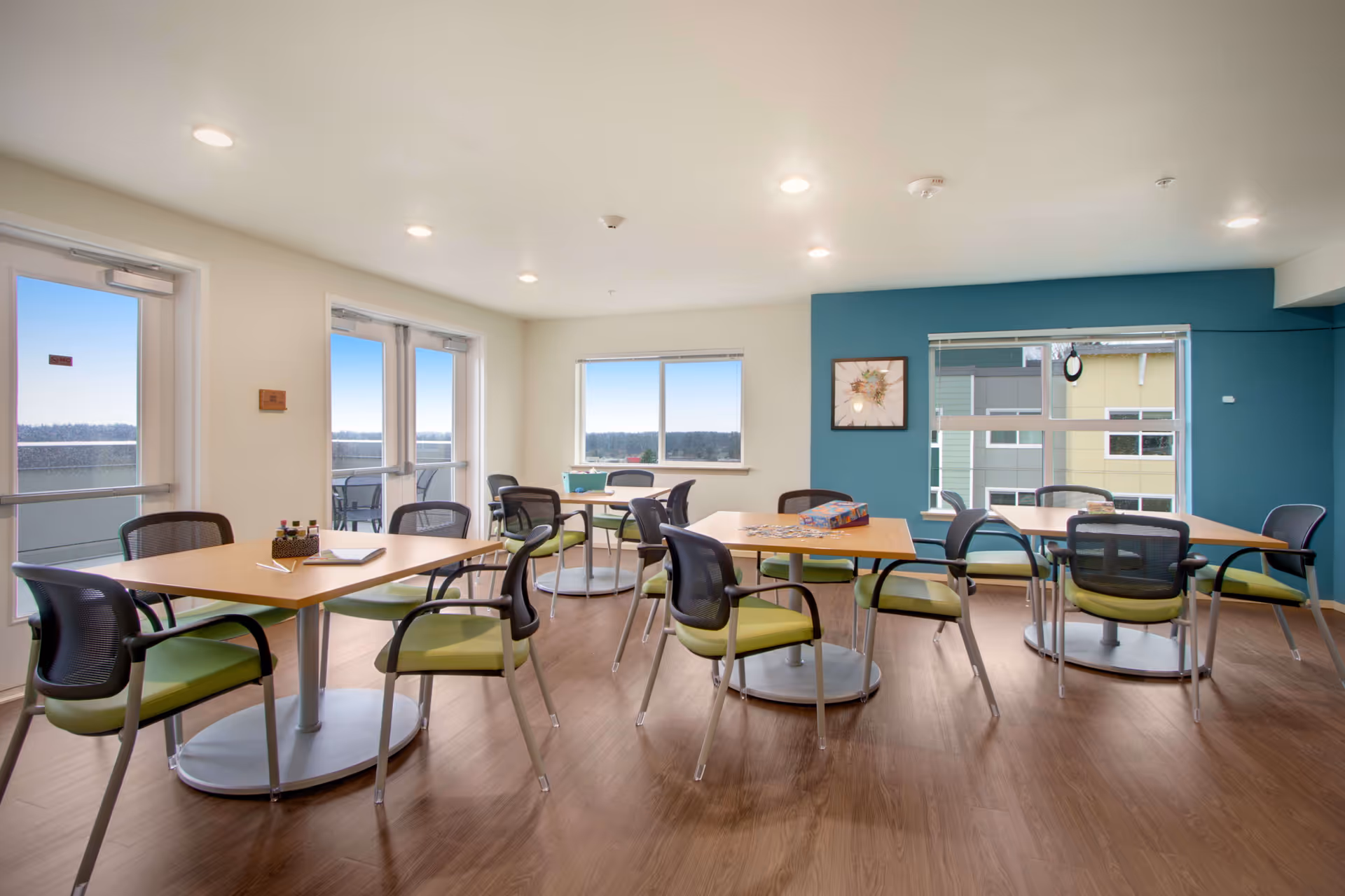 A bright and spacious common area with several square wooden tables and green cushioned chairs arranged around them. The room has large windows letting in natural light and a door leading to an outdoor balcony. The walls are painted white with one accent wall in teal, decorated with a framed artwork. The floor is wood, and there are ceiling lights evenly spaced across the ceiling.