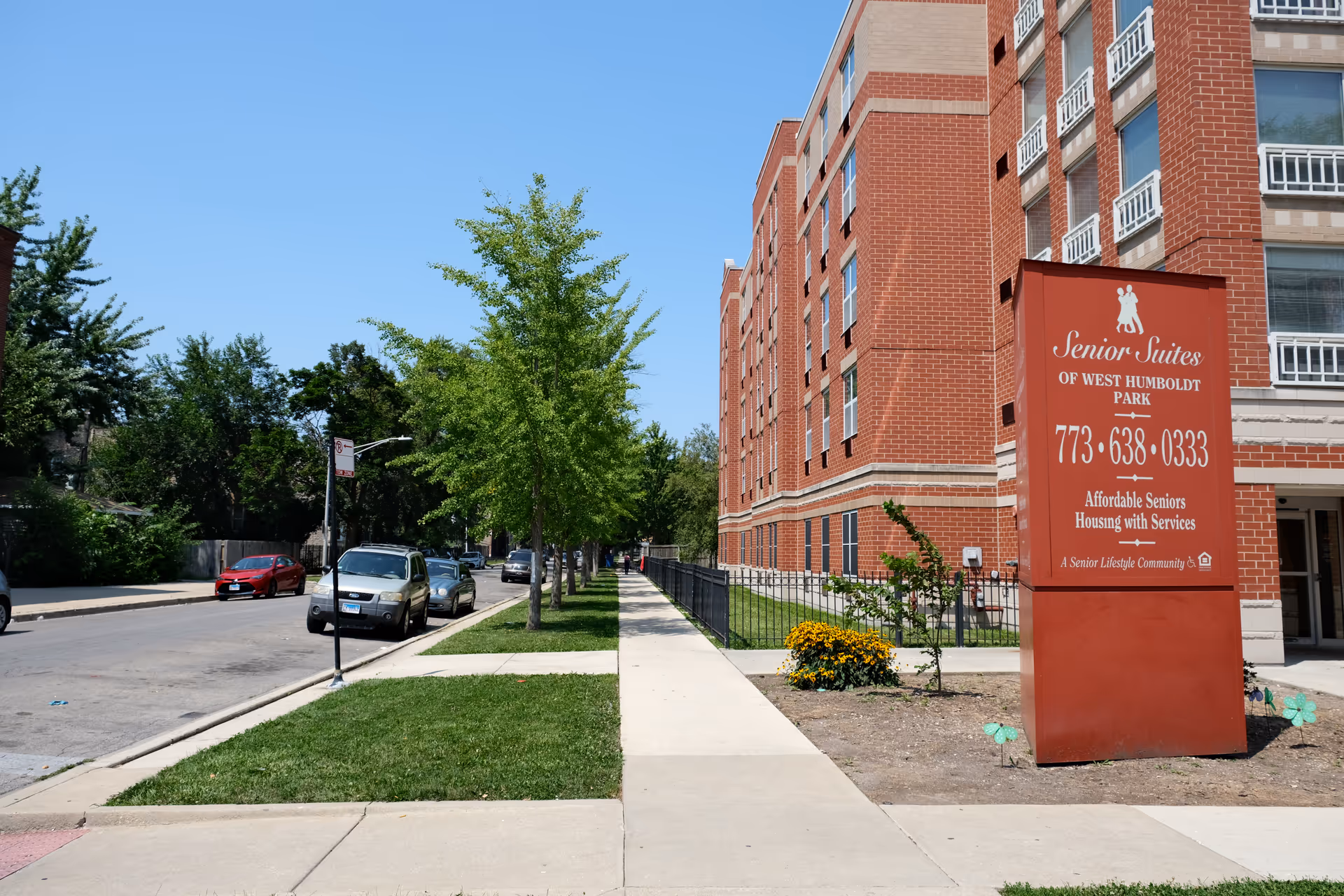 Sidewalk and street lined with trees and parked cars beside a red-brick Senior Suites of West Humboldt Park building and its large entrance sign.