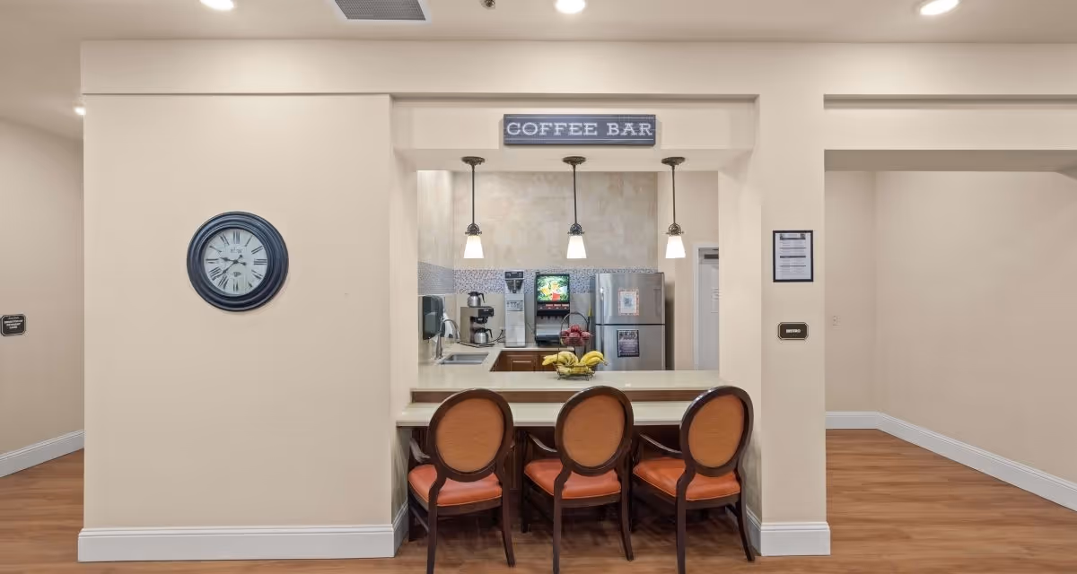 Interior view of a coffee bar area in a senior living facility with a counter and three chairs with orange cushions. Behind the counter are a coffee machine, a beverage dispenser, a refrigerator, and a sink. A wall clock is mounted on the left wall, and a sign above the counter reads 'COFFEE BAR'. The floor is wooden, and the walls are painted beige.