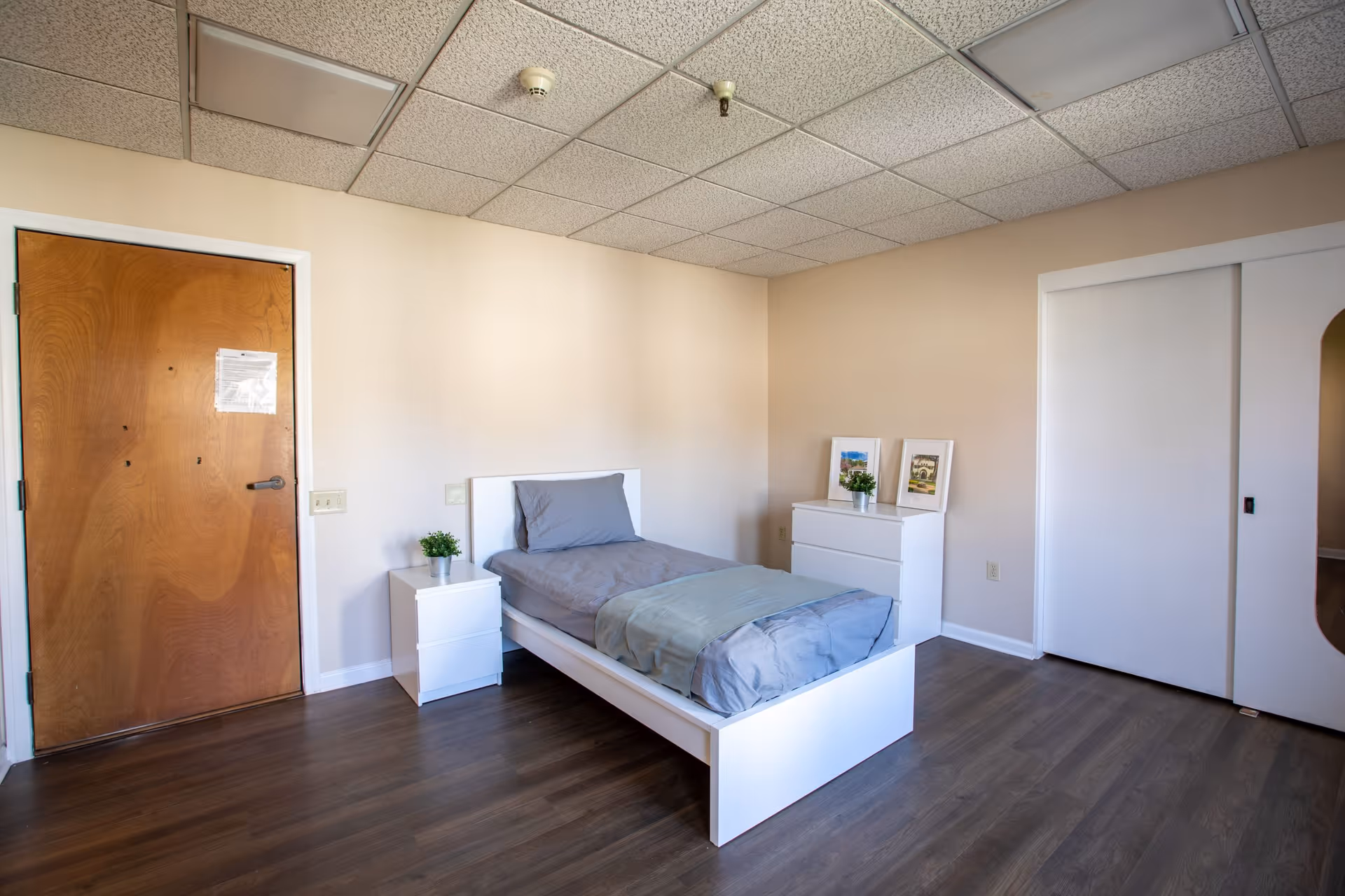 A simple bedroom with a single white bed frame and gray bedding, a white nightstand with a small potted plant, a white dresser with two framed pictures and another small potted plant, a wooden door with a paper attached, and a sliding closet door with a mirror on the right side. The room has beige walls and a dark wood floor.