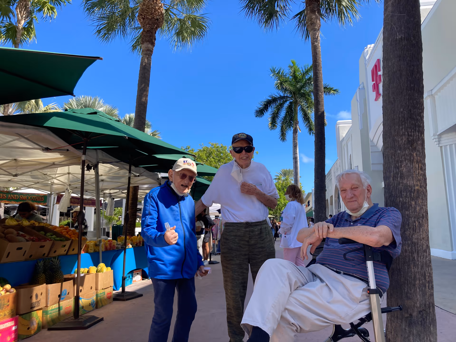 Three elderly men outdoors at a market with palm trees and blue sky in the background. Two men are standing, one wearing a blue jacket and white cap, the other in a white shirt and dark cap. The third man is sitting on a walker with a seat, wearing a striped shirt and light pants. Market stalls with fruits and green umbrellas are visible on the left side.