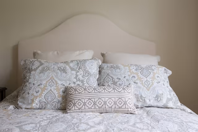 A neatly made bed with a beige upholstered headboard, two large pillows with a gray and beige paisley pattern, two plain beige pillows behind them, and a smaller rectangular pillow with a geometric pattern in front. The bedding features a matching paisley design in soft gray and beige tones.