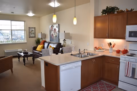 Interior view of a senior living facility apartment showing a kitchen with wooden cabinets, a white dishwasher, stove, and microwave. The kitchen counter has a double sink and pendant lights hanging above. Adjacent to the kitchen is a living area with a brown leather sofa, a coffee table, an armchair, lamps, and wall art. A large window allows natural light into the space.