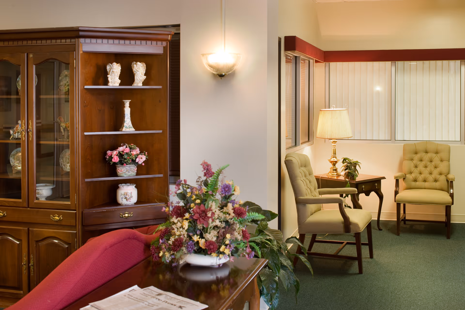 A cozy sitting area with upholstered armchairs, a side table lamp, a floral arrangement, and a wooden display cabinet.