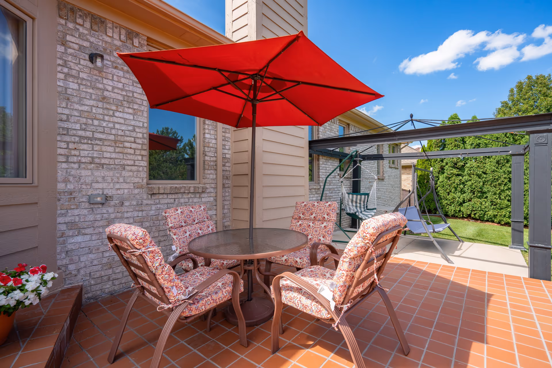 Outdoor patio area with a round glass table surrounded by four cushioned chairs with patterned fabric. A large red umbrella provides shade over the table. In the background, there is a swing set with two seats and a green lawn bordered by tall bushes under a blue sky with some clouds.