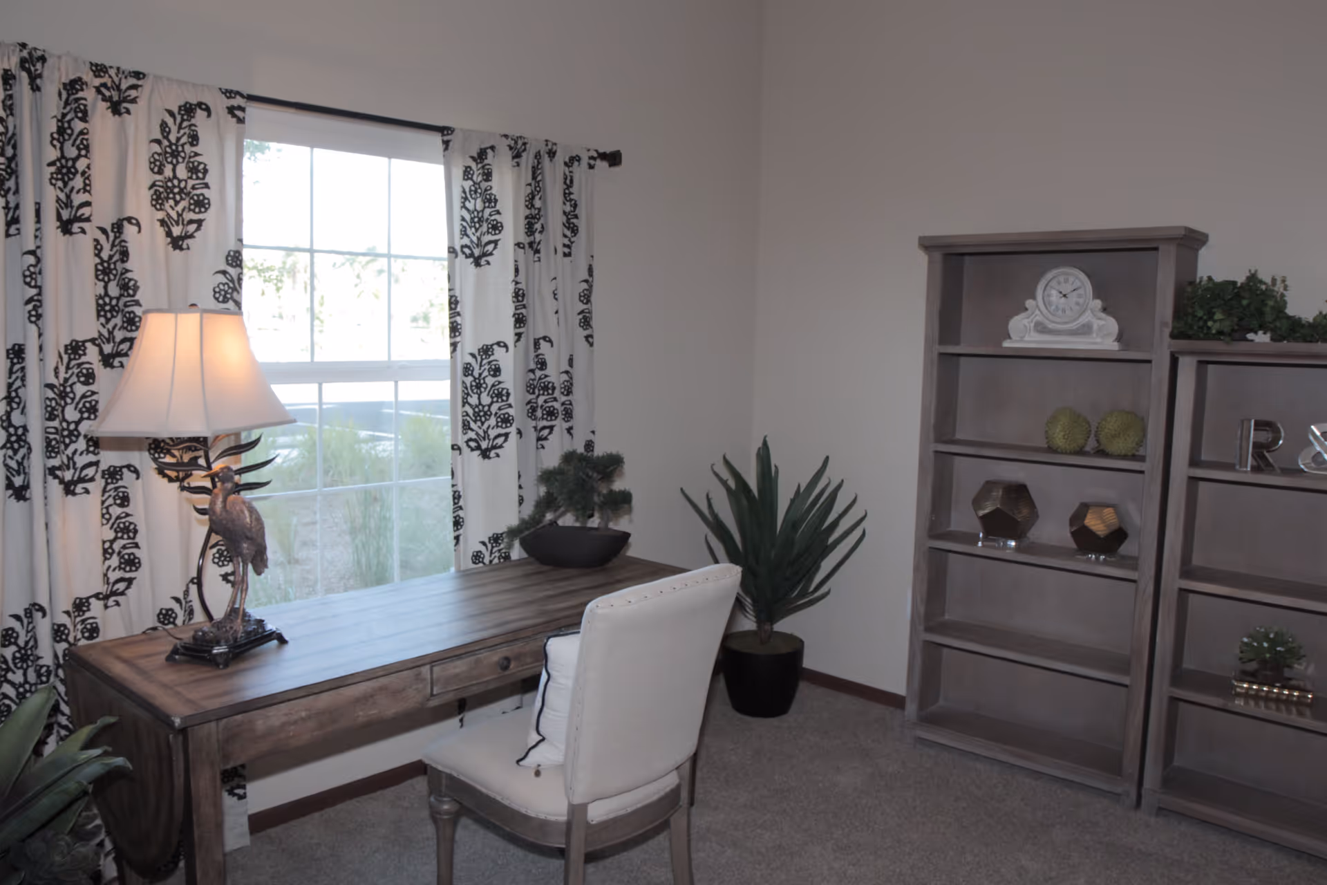 A cozy room with a wooden desk and a white cushioned chair. The desk has a decorative lamp with a bird design and a small plant. Behind the desk is a window with white curtains featuring a black floral pattern. To the right, there are two wooden bookshelves with decorative items including a clock, green spherical objects, and letters. A potted plant is placed in the corner of the room.