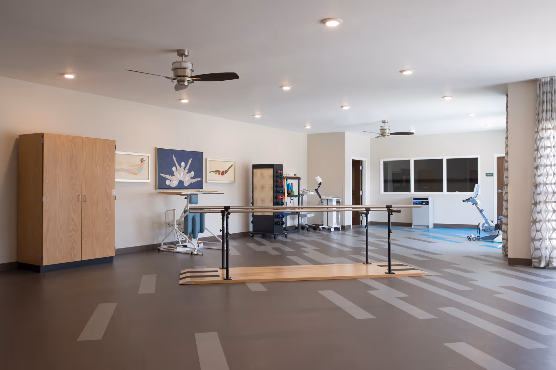 A spacious rehabilitation room with parallel bars for walking exercises in the center, exercise equipment including a stationary bike and weights, ceiling fans, and framed artwork on the walls. The room has large windows with patterned curtains allowing natural light to enter.