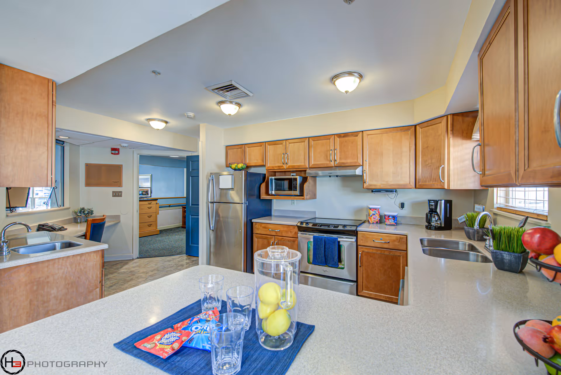A bright and clean kitchen with wooden cabinets, stainless steel refrigerator, stove, and microwave. The kitchen has two sinks, a coffee maker, and a countertop with a pitcher containing lemons, four glasses, and snack packets. There is a doorway leading to another room with blue walls and furniture.