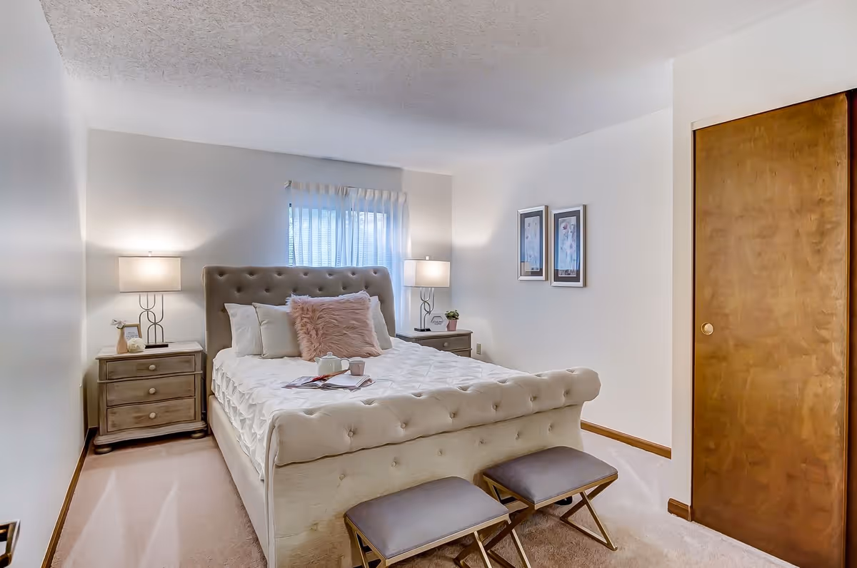 A cozy bedroom with a tufted beige bed featuring white and pink pillows, two matching nightstands with lamps on either side, a window with sheer curtains behind the bed, two framed pictures on the wall, and a wooden closet door to the right. Two small cushioned stools are placed at the foot of the bed.
