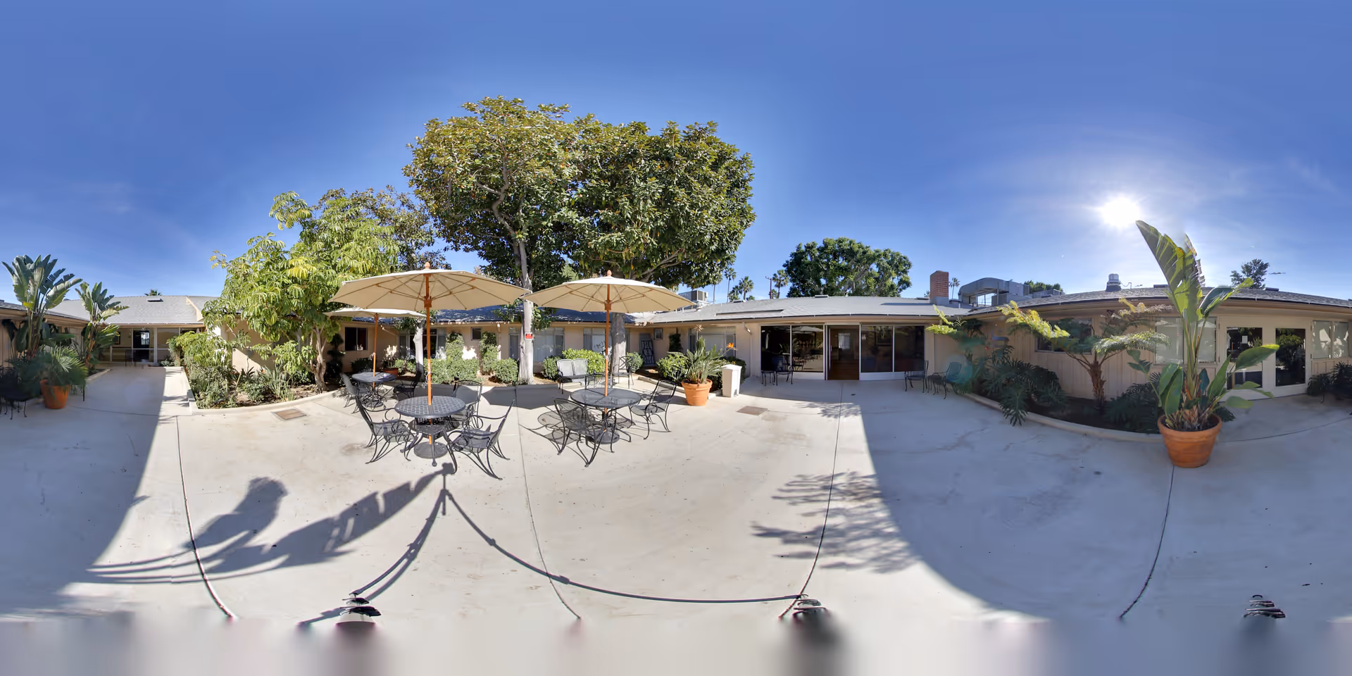Outdoor courtyard area at Advanced Rehab Center of Tustin featuring several round metal tables with chairs and large beige umbrellas providing shade. The courtyard is surrounded by single-story building walls with windows and doors, and there are various potted plants and trees around the space under a clear blue sky with bright sunlight.
