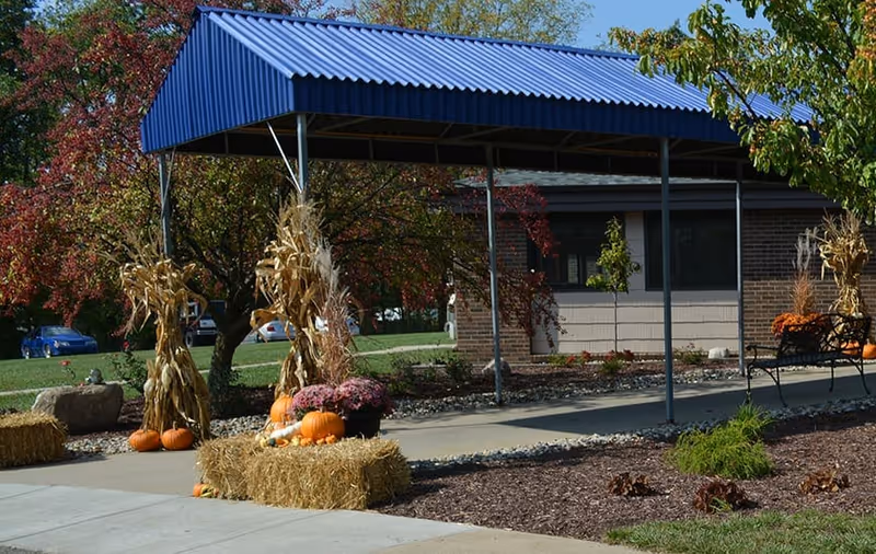 Covered entrance canopy in front of a brick building decorated with hay bales, pumpkins, mums and cornstalks for fall.