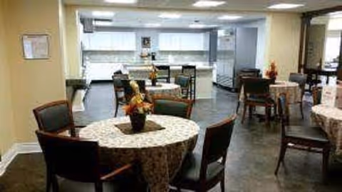 Dining area with round tables covered in patterned tablecloths, chairs, and a kitchen/service counter in the background.
