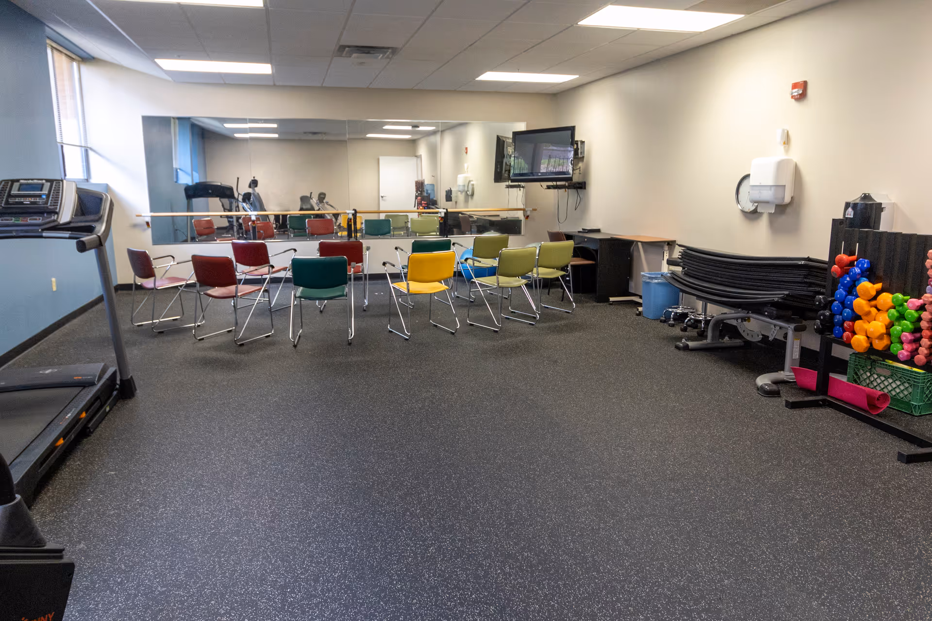 A fitness or exercise room with a treadmill on the left, colorful chairs arranged in a semicircle in the center, a large wall mirror behind the chairs, a wall-mounted TV on the right, and a rack of colorful dumbbells and exercise mats stacked on the right side.