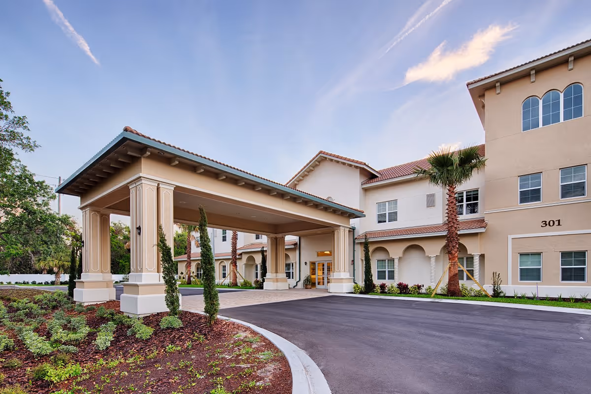 Entrance area of Lexington Place senior living facility featuring a covered drop-off with columns, landscaped garden beds with small trees and shrubs, and a multi-story beige building with arched windows and red-tiled roof under a clear blue sky.