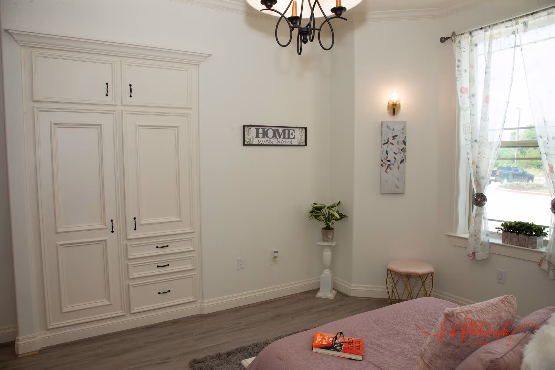 A cozy bedroom with a bed covered in a mauve blanket and pillows, a small stool with a pink cushion, a window with floral curtains, a potted plant on a white pedestal, a wall-mounted light fixture, a decorative wall sign that reads 'HOME sweet home', and a built-in white wardrobe with drawers and cabinets.