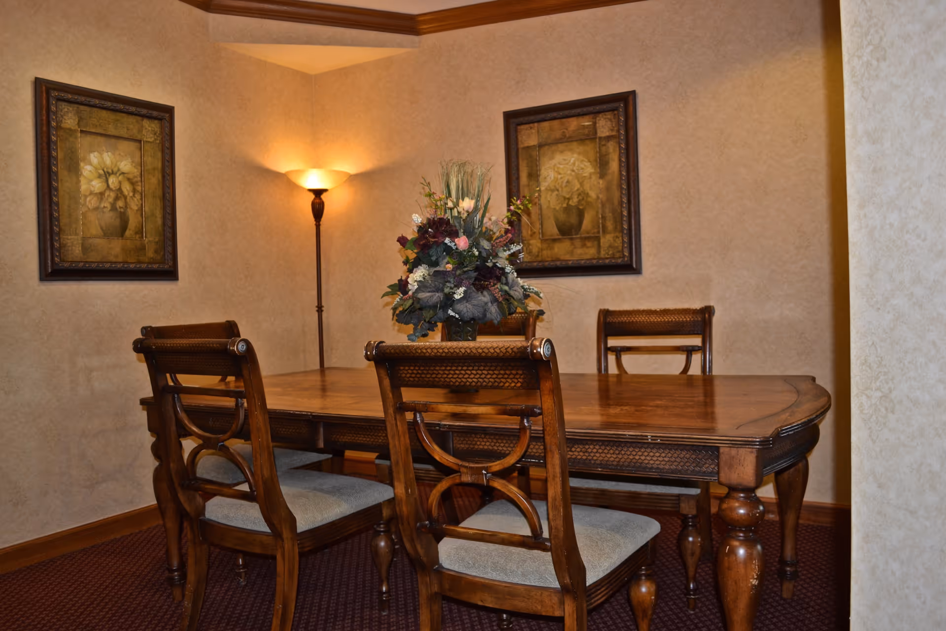 A traditional dining room with a wooden dining table and four matching chairs with cushioned seats. A floral centerpiece is placed on the table. The walls are beige with two framed floral paintings and a tall floor lamp in the corner providing warm light.