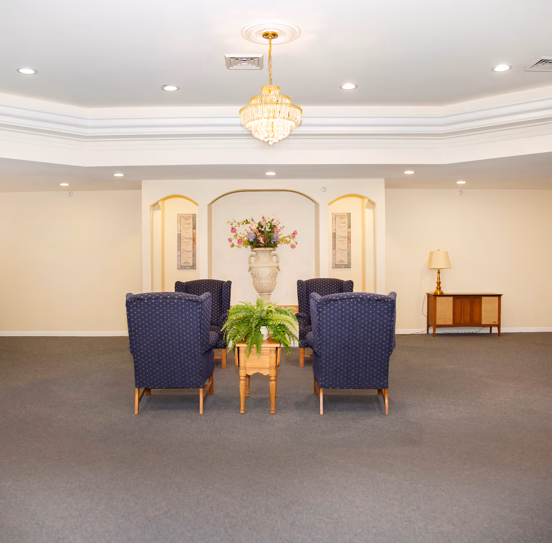 A sitting area in an assisted living facility with four blue upholstered armchairs arranged around a small wooden table with a green potted plant. Behind the chairs is a large decorative vase with flowers placed in a recessed wall niche, flanked by two framed wall art pieces. A chandelier hangs from the ceiling, and there is a wooden side table with a lamp on the right side of the room. The room has beige walls and gray carpet.