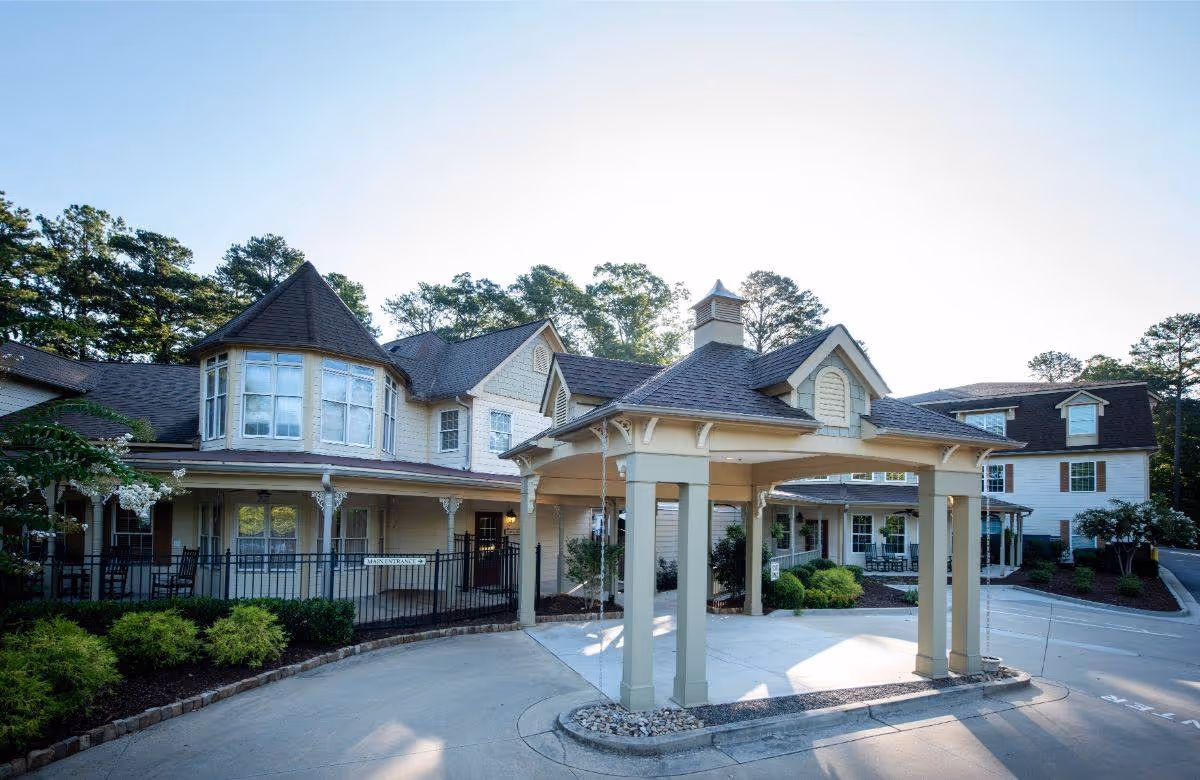 Exterior view of Highlands Senior Living Norcross showing a large covered entrance with pillars, surrounded by a multi-story building with numerous windows and a turret-like architectural feature. The area is landscaped with bushes and trees under a clear sky.