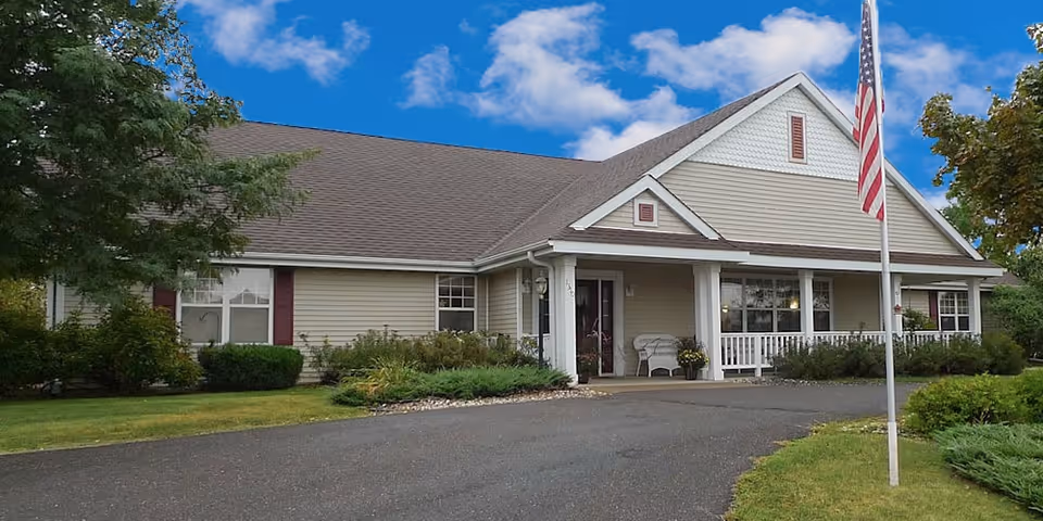 Front exterior of a single-story senior living facility with a covered porch, American flag, and landscaped lawn.