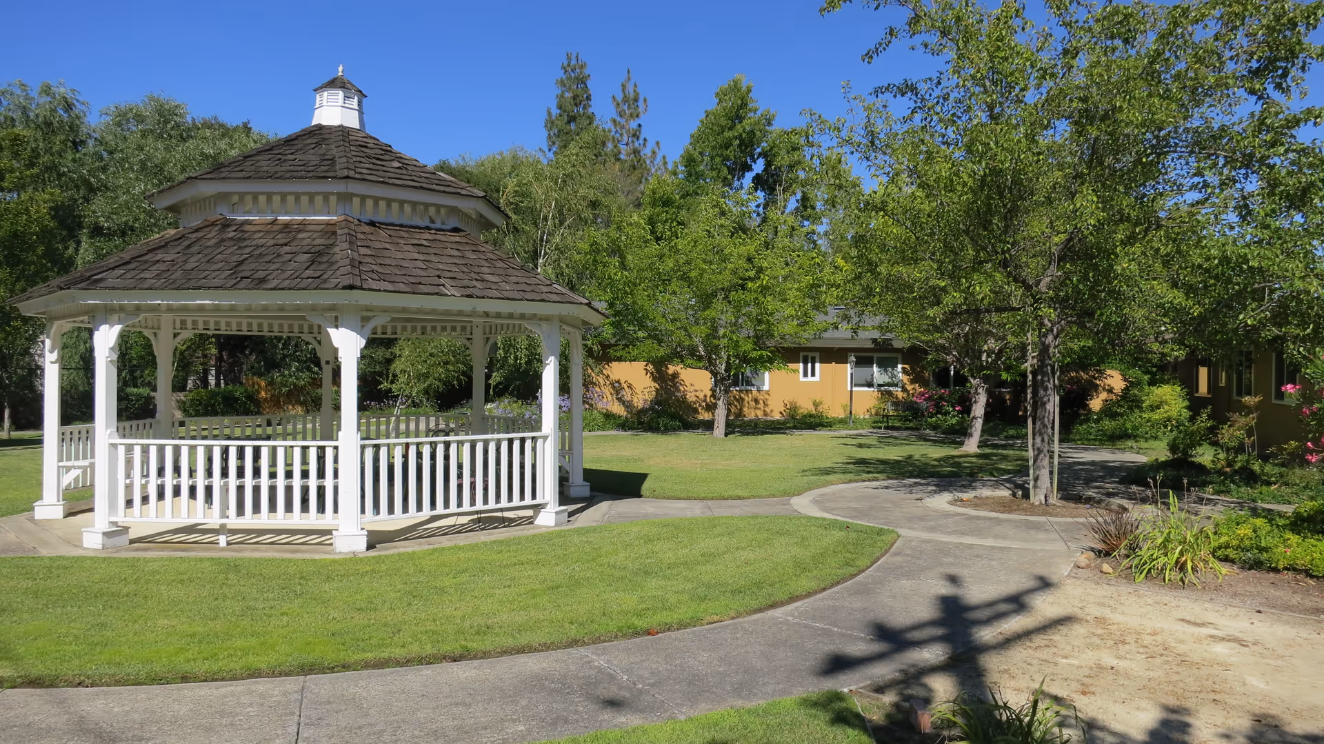 A white wooden gazebo with a shingled roof situated on a well-maintained lawn with a curved concrete pathway. Surrounding the gazebo are green trees and shrubs, with a yellow building partially visible in the background under a clear blue sky.