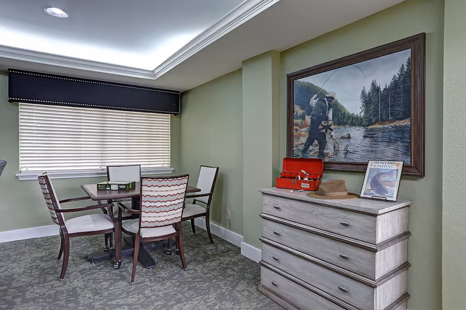 A small dining area with a wooden table and four upholstered chairs with patterned backs. There is a large window with closed blinds and a dark valance above it. To the right, there is a light-colored wooden chest of drawers with a red toolbox, a brown hat, and a framed book titled 'Hunting & Fishing' on top. Above the chest hangs a large framed painting of a person fly fishing in a river surrounded by trees.