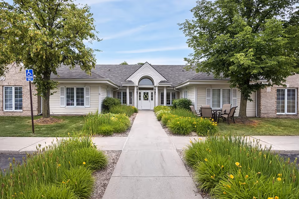 Front exterior view of a single-story senior living facility building with a central entrance featuring double doors and an arched window above. The building is surrounded by green grass, trees, and landscaped flower beds with yellow flowers. There is a concrete walkway leading to the entrance and a small outdoor seating area with a table and chairs on the right side. A handicap parking sign is visible on the left side near the building.