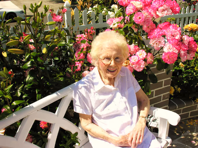 An elderly woman with white hair and glasses sitting on a white wooden bench outdoors, smiling. She is surrounded by vibrant pink and yellow flowers with green foliage and a white picket fence in the background.