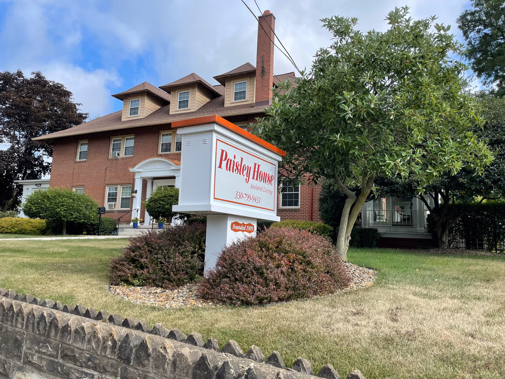 Exterior view of a large brick building with multiple windows and dormer windows on the roof. In front of the building is a white sign with red text that reads 'Paisley House Assisted Living 330-799-9431' surrounded by bushes and a tree. The sky is partly cloudy.