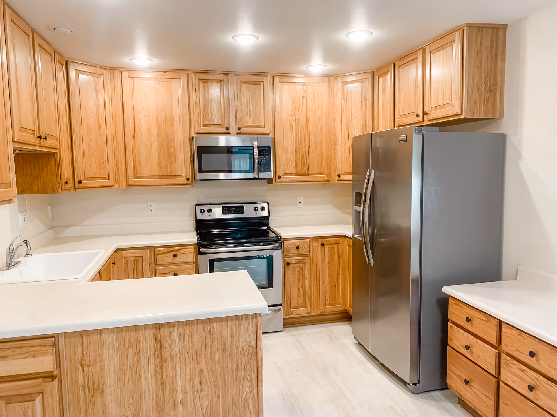 A modern kitchen with light wood cabinets, a stainless steel refrigerator, stove, and microwave. The kitchen has white countertops and recessed ceiling lights.