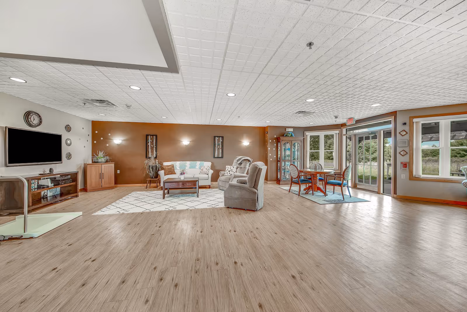 A spacious senior living facility common area with wood flooring, a white couch with a colorful throw, two gray armchairs, a wooden coffee table on a patterned rug, a TV mounted on the wall, a small dining table with four chairs near large windows and a glass door leading outside, and decorative wall art and plants.