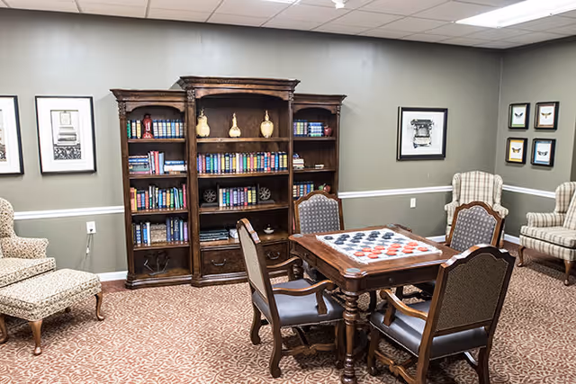 A cozy room with a wooden bookshelf filled with books and decorative items against a gray wall. In front of the bookshelf is a wooden table with a checkers game set up, surrounded by four upholstered chairs. The room also features patterned carpet, two armchairs with plaid upholstery, and framed artwork on the walls.