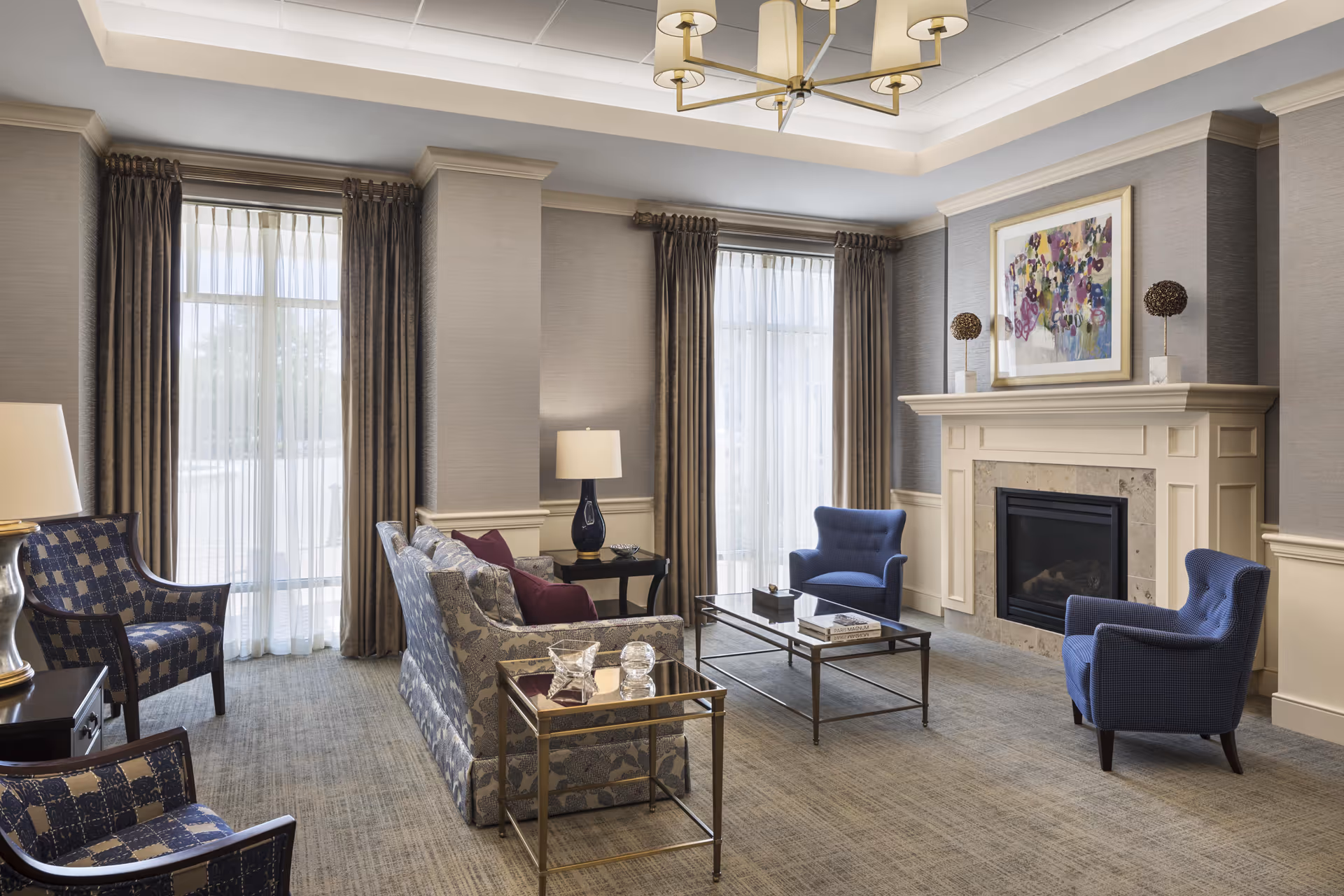 A well-lit living room with two large windows covered by sheer white curtains and brown drapes. The room features a beige carpet, a fireplace with a mantel decorated with two spherical sculptures and a colorful abstract painting above it. There are two blue armchairs, a patterned sofa with purple cushions, a glass coffee table, and side tables with lamps. The ceiling has a modern chandelier.