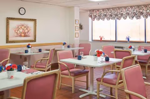 A bright dining room with pink cushioned chairs around white tables and flowers.